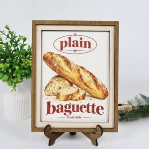 May include: A rectangular wooden frame holding a print of a golden-brown baguette. The text reads plain at the top and baguette fresh daily at the bottom in red font. The frame is supported by a dark brown wooden stand on a white surface.