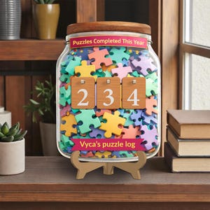 May include: A clear glass jar filled with colorful puzzle pieces, with a wooden lid and base. The jar has a label that reads "Puzzles Completed This Year" and a banner that says "Vyca's puzzle log". The jar displays the number 234.
