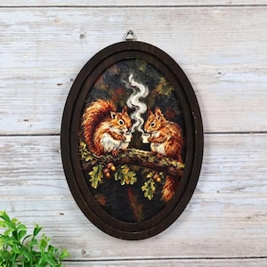 May include: An oval-framed wall art piece featuring an oil painting of two brown squirrels perched on an oak branch. Each squirrel holds a small white cup with rising steam. The frame is dark brown wood with a metal hanging loop at the top.