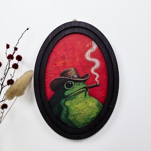 May include: An oval-framed oil painting on canvas featuring a green frog wearing a brown cowboy hat and smoking a cigar. The background is a textured, vibrant red with a thin trail of white smoke rising from the cigar into the upper area.