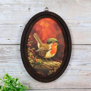 May include: An oval-shaped wall hanging featuring a painting of a small bird with a red breast and a green cap, perched on a branch with red berries against a warm orange and brown background, enclosed in a dark brown wooden frame with a metal hanger.