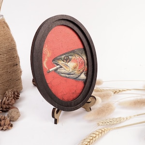 May include: An oval-shaped dark wood frame containing a canvas print of a fish with a cigarette in its mouth. The fish is rendered in shades of green, silver, and pink, set against a textured, vibrant red background with a thin trail of smoke.