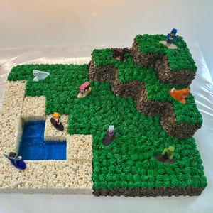 Puede incluir: Un pastel rectangular decorado para parecerse a un paisaje de videojuego. El pastel presenta glaseado verde para la hierba, glaseado marrón para los acantilados y una sustancia gelatinosa azul para el agua. Pequeñas figuras de personajes se colocan en la escena.