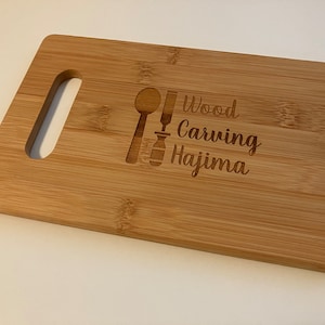 May include: A light brown bamboo cutting board with a handle. The board is engraved with a spoon and knife, and the text "Wood Carving Hajima".