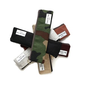 May include: A set of six different fabric pencil cases in various colors and patterns, including camouflage, black, brown, gray, and beige. Each case has a white label with the text "Independent Reign" printed on it.