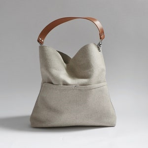 May include: A beige linen tote bag with a brown leather handle and silver hardware. The bag has a large pocket on the front.