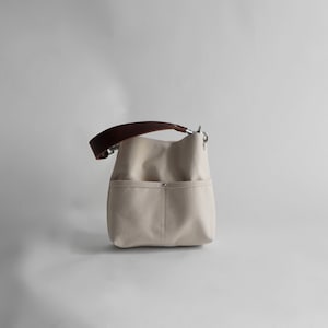 Pode incluir: Uma bolsa tote de lona bege com uma al&ccedil;a de couro marrom e ferragens prateadas. A bolsa tem um bolso grande na frente.