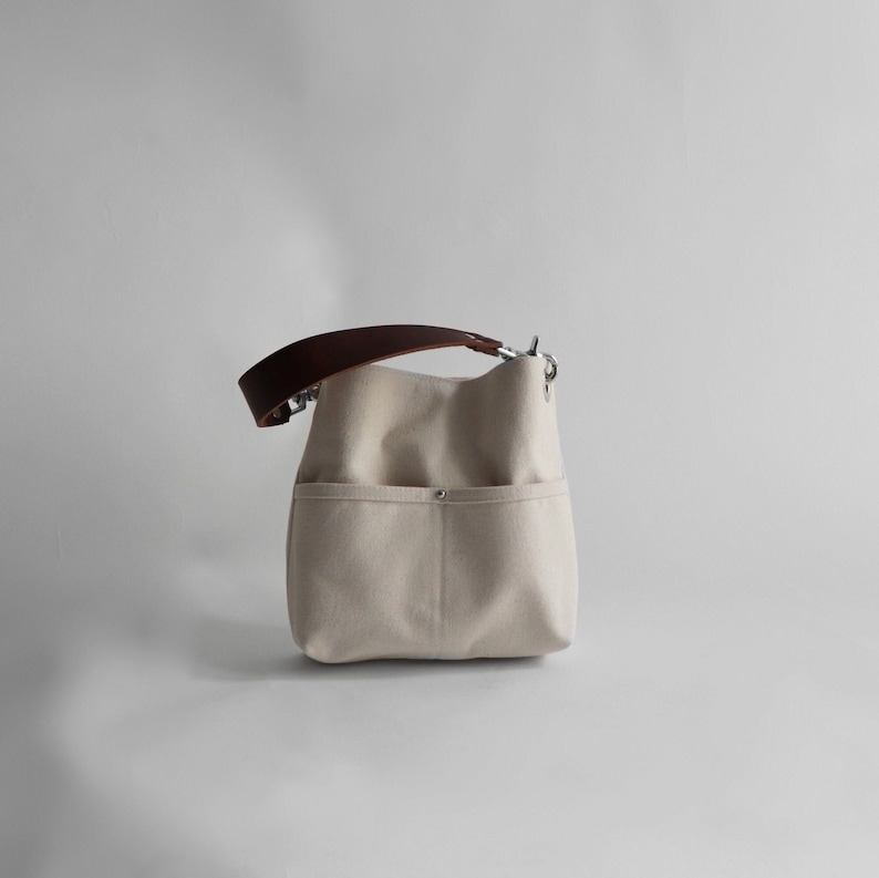 Pode incluir: Uma bolsa tote de lona bege com uma al&ccedil;a de couro marrom e ferragens prateadas. A bolsa tem um bolso grande na frente.