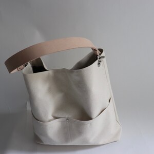 May include: A white canvas tote bag with a light brown leather strap and silver hardware. The bag has a large main compartment and two smaller pockets on the front.