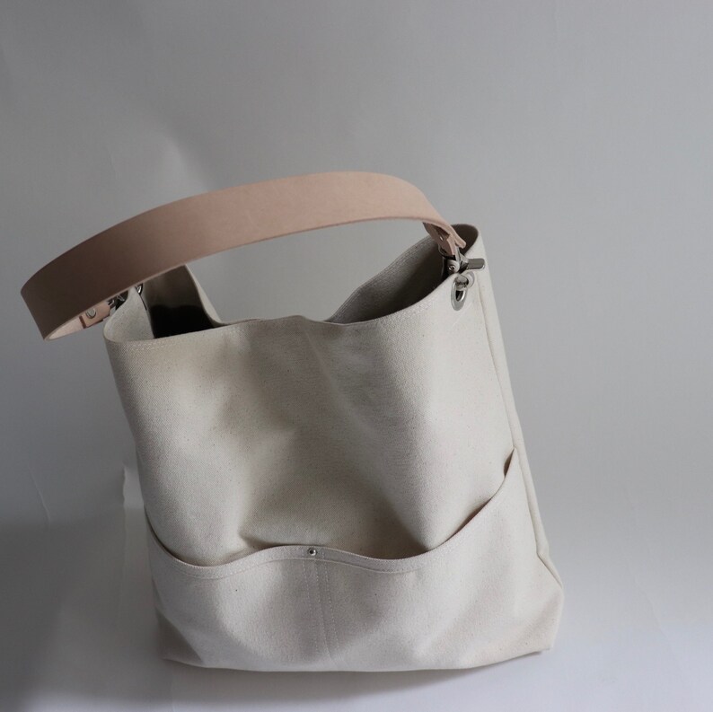 May include: A white canvas tote bag with a light brown leather strap and silver hardware. The bag has a large main compartment and two smaller pockets on the front.