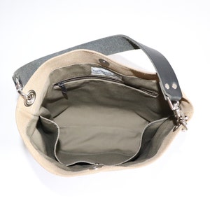 May include: A beige canvas tote bag with a black leather strap and a zippered pocket inside. The bag has a silver metal clasp and a silver metal ring on the strap.