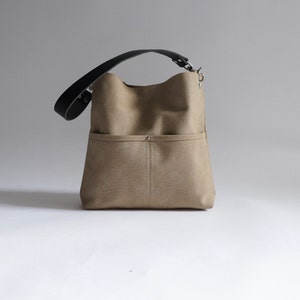 May include: A beige canvas tote bag with a black leather strap and a silver buckle. The bag has a large front pocket with a silver button.