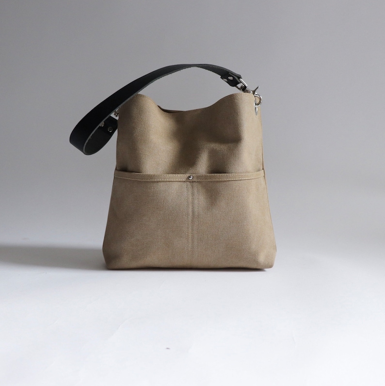 May include: A beige canvas tote bag with a black leather strap and a silver buckle. The bag has a large front pocket with a silver button.