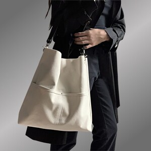 May include: A large, white canvas tote bag with a single pocket on the front. The bag has a black strap and silver hardware.