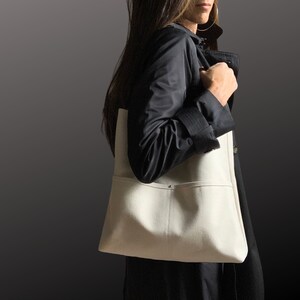 May include: A woman wearing a black jacket carries a white canvas tote bag with a single pocket on the front.