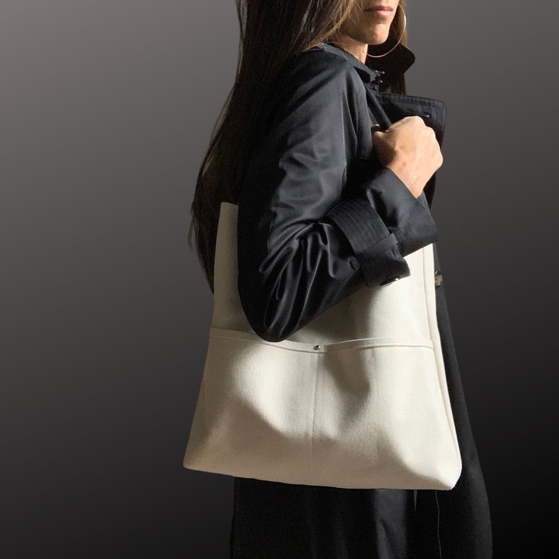 May include: A woman wearing a black jacket carries a white canvas tote bag with a single pocket on the front.