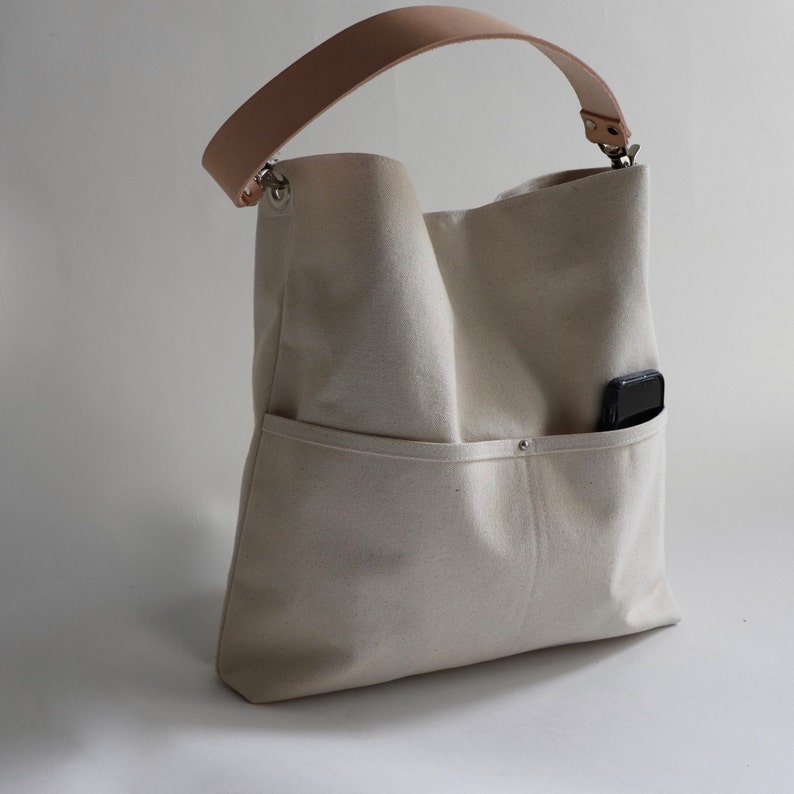 May include: A white canvas tote bag with a brown leather strap and a small pocket on the side. The bag has a zipper closure and a silver metal buckle.