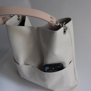 May include: A white canvas tote bag with a light brown leather strap and a silver metal clasp. The bag has a pocket on the front with a black phone inside. The phone screen shows the time 10:47.