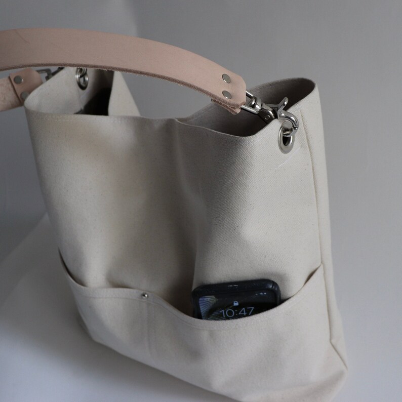 May include: A white canvas tote bag with a light brown leather strap and a silver metal clasp. The bag has a pocket on the front with a black phone inside. The phone screen shows the time 10:47.