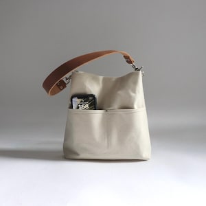 May include: A beige canvas tote bag with a brown leather strap and a zipper closure. A cell phone with a digital clock displaying 1:58 is in the front pocket.