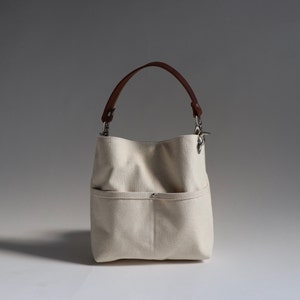 Pode incluir: Uma bolsa tote de lona branca com al&ccedil;a de couro marrom e fecho de prata. A bolsa tem um bolso grande na frente.