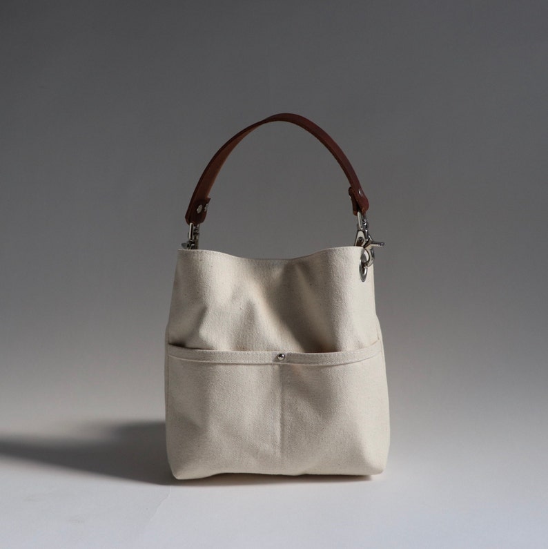 Pode incluir: Uma bolsa tote de lona branca com al&ccedil;a de couro marrom e fecho de prata. A bolsa tem um bolso grande na frente.