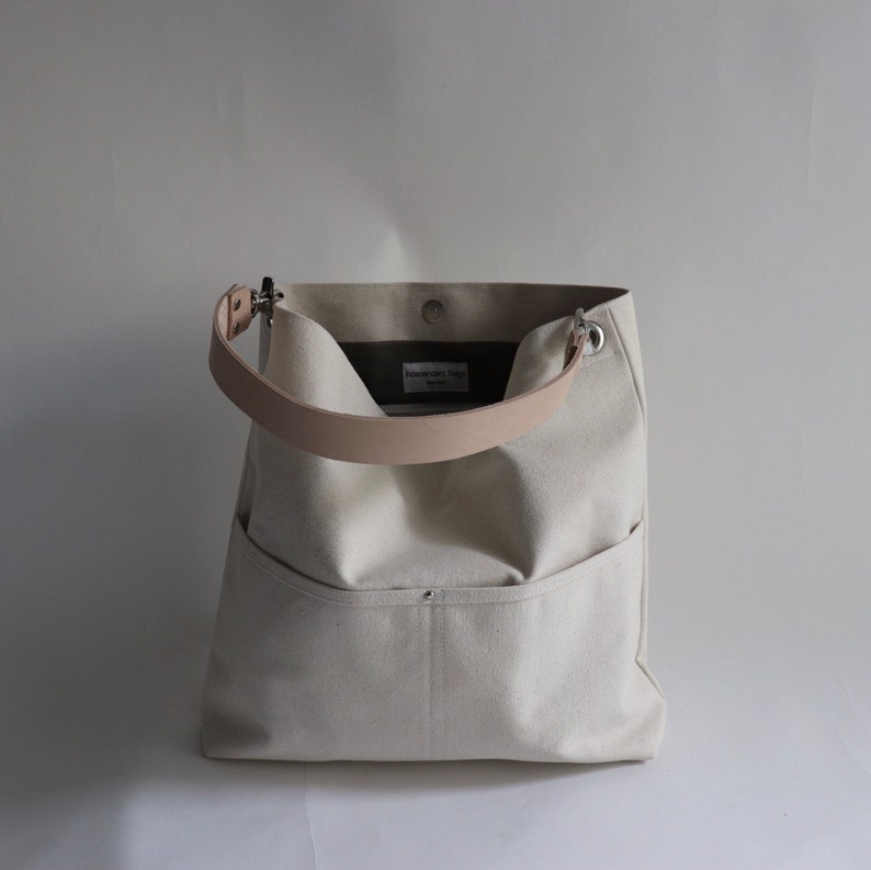 May include: A white canvas tote bag with a light brown leather strap and a small pocket on the front. The bag has a label that says "Housewares Design" on it.