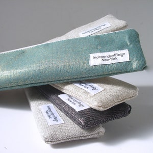 May include: A stack of four pencil cases in different colors and fabrics. The top case is teal with gold glitter, the second is beige linen, the third is dark gray, and the bottom is beige linen. Each case has a white label with the text "IndependentReign New York".
