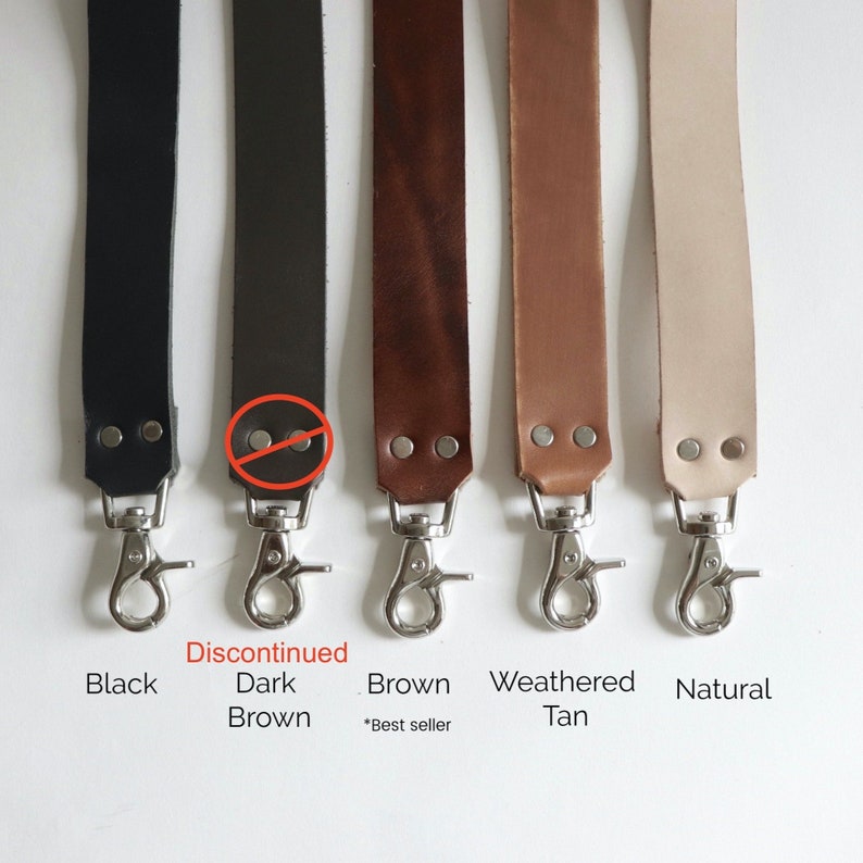 May include: Five leather camera straps in different colors: black, dark brown, brown, weathered tan, and natural. The straps have silver metal clasps and are shown against a white background. The dark brown strap is marked as discontinued.