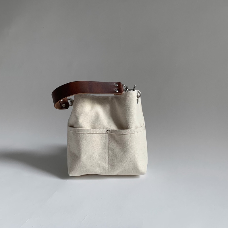 Pode incluir: Uma bolsa tote de lona branca com uma al&ccedil;a de couro marrom e ferragens prateadas. A bolsa tem um bolso grande na frente e um bolso menor no lado.