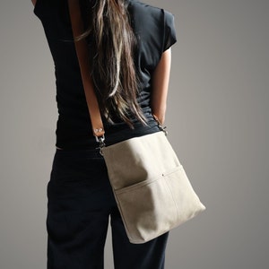 May include: A beige canvas crossbody bag with a brown leather strap and a silver buckle. The bag has a large pocket on the front.