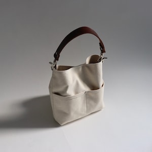 Pode incluir: Uma bolsa tote de lona branca com al&ccedil;as de couro marrom e ferragens prateadas. A bolsa tem dois bolsos grandes na frente.