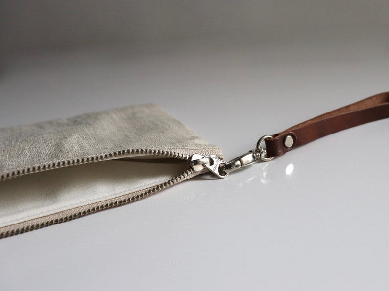May include: A beige linen zippered pouch with a brown leather strap and a silver clasp.