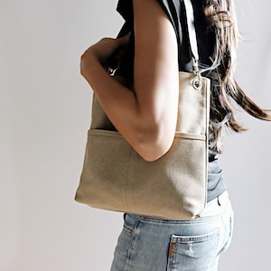 May include: A beige canvas tote bag with a long strap and a front pocket. The bag is worn over the shoulder of a person wearing a black shirt and blue jeans.