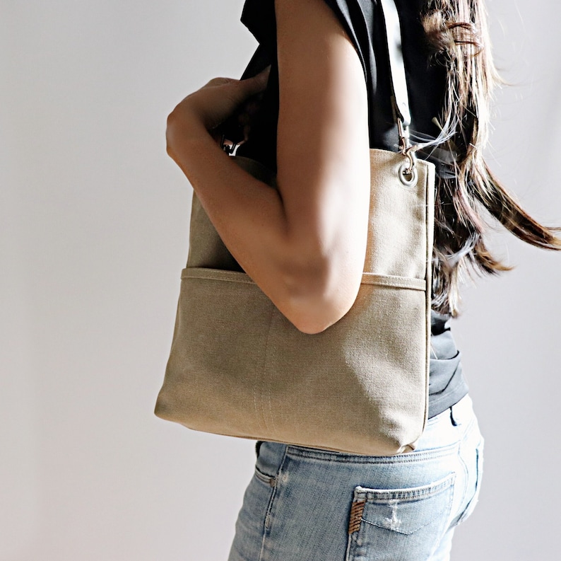 May include: A beige canvas tote bag with a long strap and a front pocket. The bag is worn over the shoulder of a person wearing a black shirt and blue jeans.