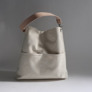 May include: A white canvas tote bag with a light brown leather strap and a silver clasp. The bag has a large pocket on the front.