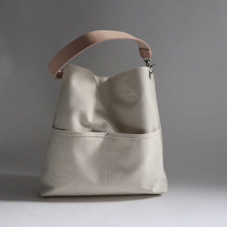 May include: A white canvas tote bag with a light brown leather strap and a silver clasp. The bag has a large pocket on the front.
