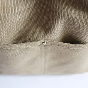 May include: Close-up of a beige canvas tote bag with a small silver rivet on the pocket.