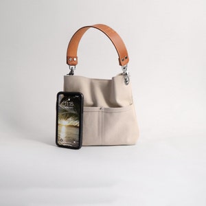 Pode incluir: Uma bolsa tote de lona bege com al&ccedil;a de couro marrom e ferragens prateadas. Uma capa de telefone preta com uma tela que mostra a hora 11:15 est&aacute; no bolso frontal da bolsa.