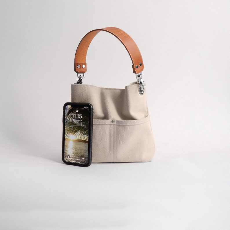 Pode incluir: Uma bolsa tote de lona bege com al&ccedil;a de couro marrom e ferragens prateadas. Uma capa de telefone preta com uma tela que mostra a hora 11:15 est&aacute; no bolso frontal da bolsa.