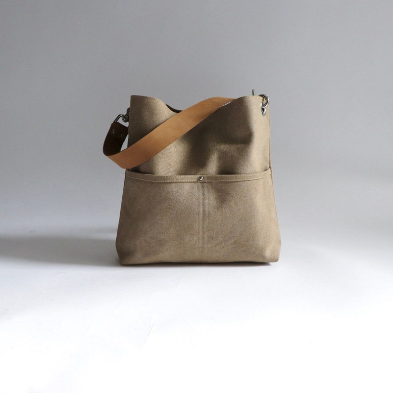May include: A beige canvas tote bag with a brown leather strap and a silver metal buckle. The bag has a large pocket on the front.