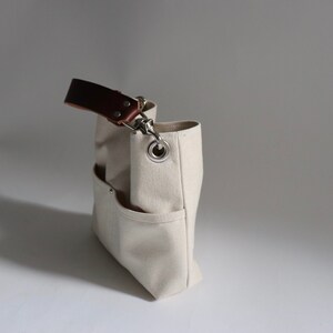Pode incluir: Uma bolsa tote de lona bege com uma al&ccedil;a de couro marrom e um anel de metal prateado. A bolsa tem v&aacute;rios bolsos e um fecho de correr.