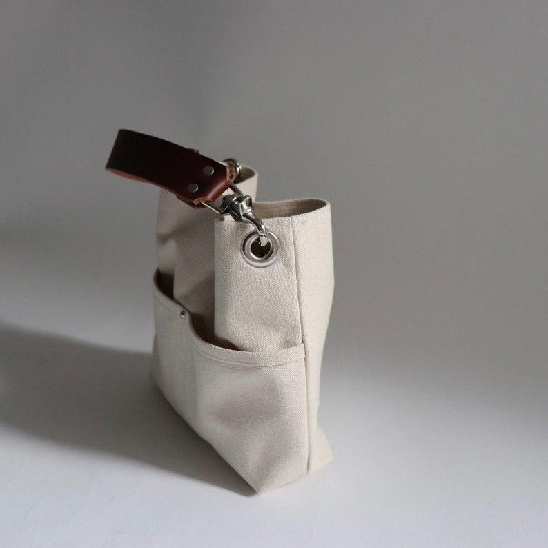 Pode incluir: Uma bolsa tote de lona bege com uma al&ccedil;a de couro marrom e um anel de metal prateado. A bolsa tem v&aacute;rios bolsos e um fecho de correr.