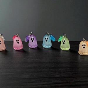 Puede incluir: Seis llaveros con forma de fantasma de colores rosa, morado, azul y verde. Cada fantasma tiene una cara negra y un pequeño lazo para sujetar las llaves. Los llaveros están colocados sobre una superficie oscura.