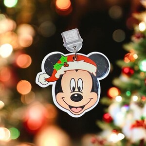 May include: A red and white Christmas bauble featuring Mickey Mouse wearing a Santa hat. The bauble is hanging from a clear plastic hook.