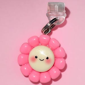 May include: A pink flower charm with a smiling yellow face and rosy cheeks. The charm is attached to a clear plastic clip.