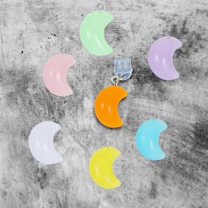 May include: Seven crescent moon shaped charms in various pastel colors, including light green, pink, lavender, white, light blue, and yellow. The charms are made of a translucent material and have a silver loop at the top.