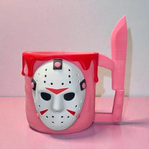 May include: A pink mug featuring a white hockey mask with red accents on the front. The top rim of the mug has a red dripping paint effect. The handle is shaped like a large pink knife, extending vertically from the side of the mug body.