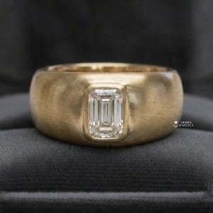 May include: A gold ring with a brushed finish, featuring a rectangular emerald-cut diamond. The ring has a wide band and a bezel setting for the diamond. The ring is displayed on a dark gray surface.