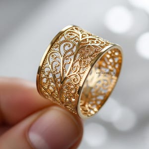 以下が含まれることがあります： 精巧な透かし彫りのデザインが施されたゴールドのフィリグリーリング。リングは、詳細な花柄のワイドバンドが特徴です。リングは手で持たれており、その繊細な職人技を披露しています。リングは存在感のあるジュエリーです。